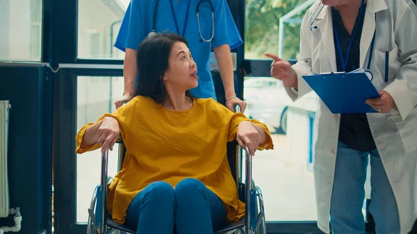 Asian woman with physical disability receiving help from nurse, having checkup visit appointment with physician. Assistant giving support to patient dealing with chronic impairment condition.