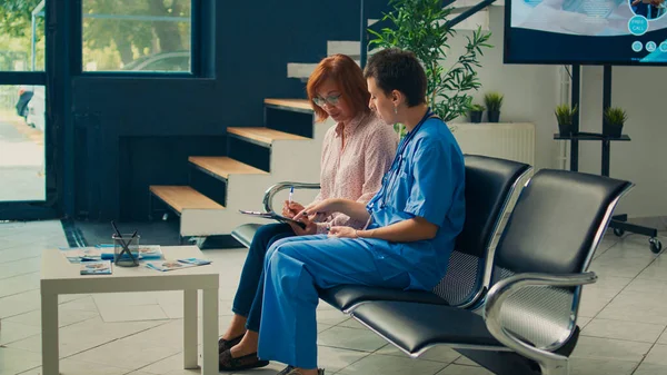 Tıp asistanı Asyalı kadının sağlık raporunu doldurmasına yardım ediyor, muayene kağıtları yazıyor ve hastane lobisinde bekliyor. Hasta klinikteki hemşireye danışmaya hazırlanıyor..