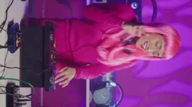 Vertical video: Cheerful performer standing at dj turntables mixing sounds during electronic performance in nightclub. Asian musician playing stereo sounds with electronics, dancing and having fun