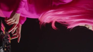 Vertical video: Asian dj having fun mixing eletronic music with techno using professional turntables, standing at dj table in club at night. Musical performer with pink hair and blouse enjoying