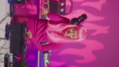 Vertical video: Artist standing at dj table performing electronic song using professional mixer console, enjoying playing music during night time in club. Perfomer with pink hair doing performance