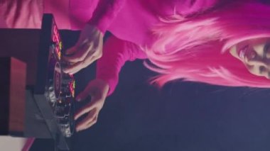 Vertical video: Smiling artist working as dj playing at mixer console, mixing techno sound with eletronic using professional audio equipment. Musician with pink hair having fun while performing in
