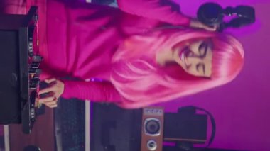 Vertical video: Musician with pink hair playing eletronic song at mixer console, listening music into headset. Asian dj perfomer enjoying mixing sounds, dancing with fans during night time in club.