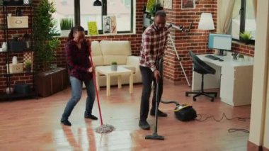 African american couple dancing and cleaning apartment rooms, using mop to wash dirt and vacuum to clean floors. Young life partners enjoying spring cleaning with washing solution.