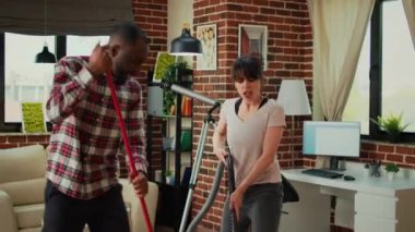 Happy life partners using tools to clean apartment, listening to music and dancing while they use mop and vacuum. Mixed race couple cleaning living room floors, sweeping dust with cleaner.