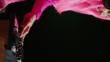 Vertical video: Artist with pink hair mixing and mastering electronic sound using professional turntables, performing music in nightclub. Dj performer playing techno song, having fun during