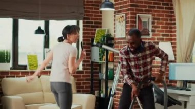 Interracial partners having fun cleaning room with vacuum cleaner and mop, dancing and listening to music. Modern cheerful people enjoying spring cleaning, vacuuming apartment floors.