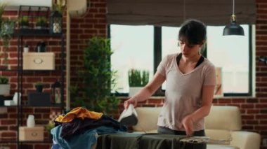Young adult using steam iron to smooth out wrinkles on clothing, ironing pile of laundered clothes on ironing table. Casual person doing spring cleaning at home, housekeeping work.