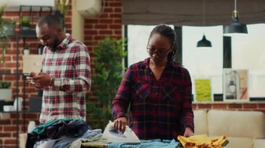 Young modern woman hitting partner with shirt after using iron, ironing washed clothes and doing household chores. Frustrated tired housewife working with laundry, spring cleaning.