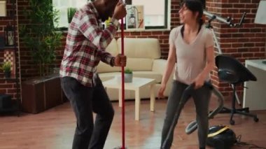 Smiling young people using tools to clean household, listening to music and dancing while they use mop and vacuum. Diverse couple cleaning apartment floors, sweeping dust with solution.