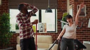 Interracial couple showing dance moves and having fun spring cleaning, mopping and vacuuming wooden floors in living room. Happy life partners enjoying using mop and vacuum cleaner.