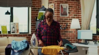 Young modern housewife using heat iron and ironing table, smoothing out creases on clothes. Female person doing household chores for spring cleaning, using laundered clothing.
