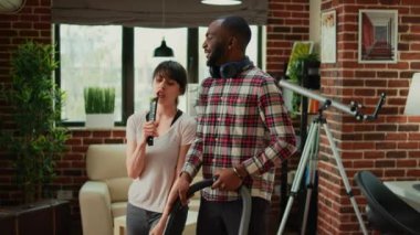 Mixed race husband and wife dancing and sweeping floors, using vacuum cleaner and all purpose cleaner. Happy man and woman laughing and enjoying spring cleaning at home, household chores.