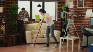 Young couple washing wooden floor with mop, husband helping wife to clean under the couch. Man and woman doing spring cleaning in living room, using all purpose cleaner and washing solution.