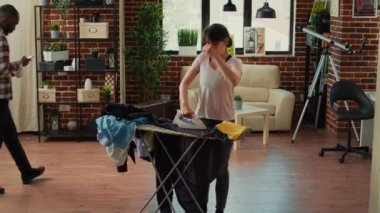 Aggressive woman ironing shirt and getting frustrated at boyfriend, needing help with house chores. Stressed exhausted girlfriend cleaning household and doing housekeeping without man.