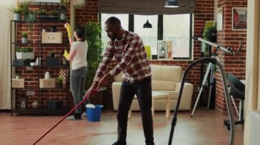 Interracial couple vacuuming floors and cleaning furniture on shelves, sweeping dust or dirt with tools. Life partners using vacuum cleaner with hose, gloves and rags to clean household. House chores.