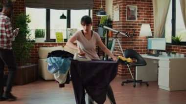Positive person singing and having fun ironing clothes in living room, girlfriend listening to music and dancing at home. Happy woman doing household chores and using iron with steam.
