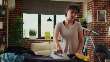 Cheerful woman dancing and having fun ironing clothes in living room, girlfriend listening to music and singing at home. Happy adult doing housework and using iron with steam after laundry.