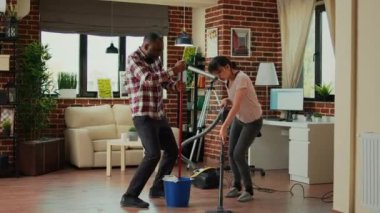 Diverse couple showing dance moves and having fun cleaning apartment, using mop to sweep dust and vacuum at home. Life partners dancing and singing, using washing solution, spring cleaning.