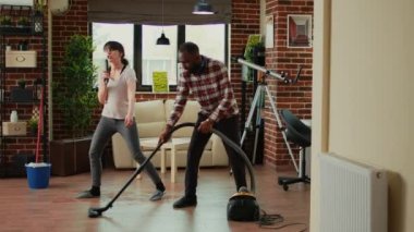 Funny cheerful people singing song and cleaning living room floors, using mop and vacuum cleaner at home. Diverse couple dancing and listening to music, washing dust and mess laughing.