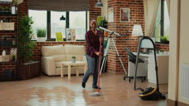 Happy woman dancing in living room and using mop to clean floors, listening to music and having fun spring cleaning. Positive housewife singing, mopping tiles and using washing solution.