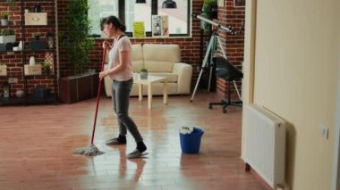 Caucasian wife cleaning apartment floors with mop, listening to music and doing chores. Cheerful woman dancing and washing dirt, doing housework in living room. Spring cleaning.