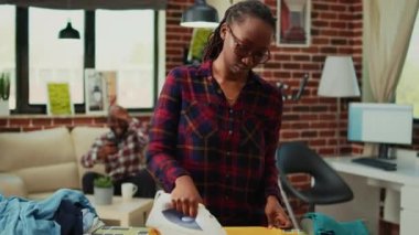 Frustrated sleepy young adult ironing clothes, getting angry at boyfriend not helping with housework. Exhausted stressed girlfriend taking care of household by herself, chores.