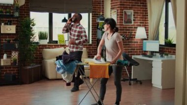 Young life partners having argument in living room, fighting over unfinished chores and spring cleaning. Aggressive woman being angry with husband while she irons clothes on ironing board.