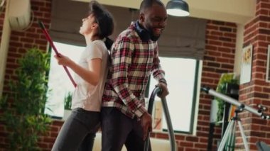 Mixed race couple vacuuming wooden floors and showing dance moves at home, having fun doing chores. Cheerful modern man and woman dancing and singing on music, acting funny. Handheld shot.