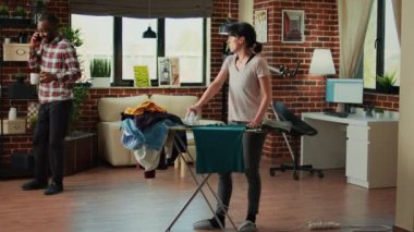 Mixed race couple having argument at home, fighting over unfinished chores and spring cleaning. Frustrated woman being angry with african american husband while she irons clothes on ironing board.