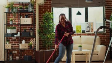 Modern smiling girlfriend listening to music and mopping wooden floors, using mop and solution in bucket. Cheerful person dancing and singing, having fun spring cleaning. Tripod shot.