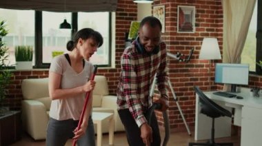 Diverse life partners dancing and sweeping dust off floors, using vacuum cleaner and washing solution. Cheerful couple laughing and enjoying spring cleaning in apartment, household chores.