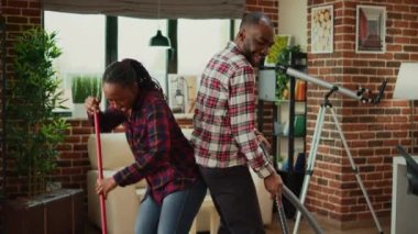 Cheerful people showing dance moves and doing spring cleaning, having fun with music while they clean wooden floors with mop and vacuum cleaner. Young couple smiling and doing chores.