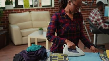 Young girlfriend enjoying doing ironing work at home, having fun listening to music and ironing laundered clothes. Cheerful woman dancing and singing while she does chores, cleaning.
