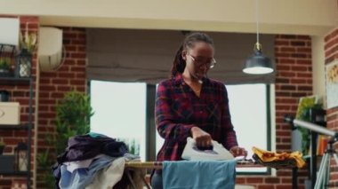 Young housewife enjoying ironing blouses in apartment, listening to music and dancing while she does chores. Modern person showing dance moves and being happy, having fun. Handheld shot.