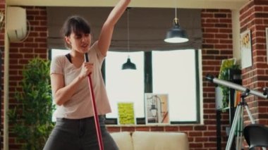 Young woman singing and dancing in living room, washing apartment floors with mop and washing solution. Cheerful housewife mopping tiles and having fun spring cleaning. Handheld shot.