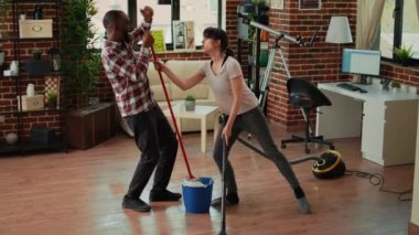 Happy man and woman doing house chores and dancing, listening to music for spring cleaning work. Cheerful couple sweeping floors and using vacuum, singing together in living room.