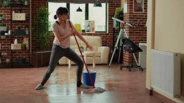 Happy funny person mopping floors with all purpose cleaner and dancing, spring cleaning. Young cheerful housewife using mop to sweep dirt and clean mess, listening to music at home.