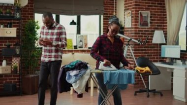 Stressed young person ironing laundered apparel while relaxed husband uses phone on couch. Exhausted and frustrated girlfriend doing housework and chores, laundry work in living room.