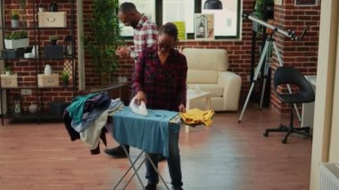 Tired modern wife ironing laundered clothes while relaxed husband uses smartphone. Disappointed and frustrated wife doing housework and chores alone, laundry work in living room.