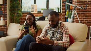 Neşeli insanlar evlerinde rahat oturuyorlar, cep telefonlarındaki sosyal medya uygulamalarına bakıyorlar ve dinleniyorlar. Erkek arkadaş ve kız arkadaş akıllı telefonu kanepede samimi bir şekilde kullanıyor..