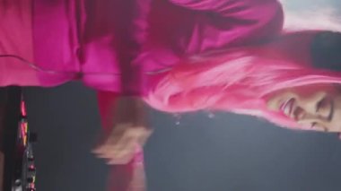 Vertical video: Performer mixing electronic sound with techno, enjoying party with fans, standing at dj table performing at party in club at night. Asian musician with pink hair performing music using