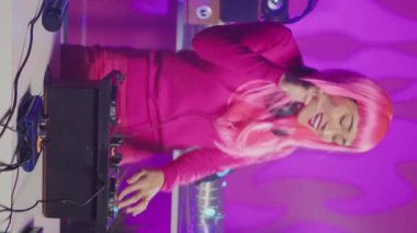 Vertical video: Smiling musician working as dj playing at turntables, mixing eletronic sound with techno using professional audio equipment. Asian artist with pink hair having fun performing in club