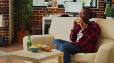 Smiling adult eating hamburger with fries and alcohol, watching favorite action movie by herself. Modern woman feeling cheerful with fast food delivery, watch show on television.
