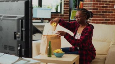 Young adult eating cheeseburgers with fries and beer, having fun with comedy movie on television. Relaxed girl enjoying cheeseburger and snacks, binge watching show in living room.