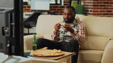 Young male adult eating slice of pizza at tv, laughing in living room on couch. Happy relaxed person enjoying takeaway meal and binge watching favorite tv show, having fun at home.