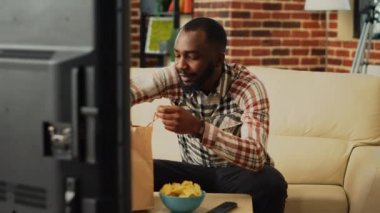 Cheerful person taking bite of cheeseburger on couch, eating burger with fries and bottle of beer. Young guy watching television and having dinner, feeling relaxed with delivery food.