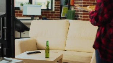 Smiling girl preparing to eat slice of pizza at tv, sitting on couch and watching comedy movie. Cheerful modern woman eating dinner from fast food meal delivery, drinking alcohol at home.