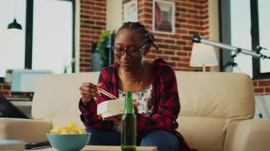 Young adult eating noodles in fast food delivery box, using chopsticks to eat asian meal and watch comedy film on tv. Girl enjoying dinner from takeout place, having fun at home.