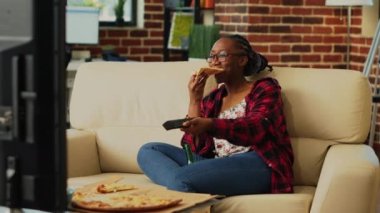 Happy woman eating pizza from delivery at home, enjoying movie on television with beer bottles and fast food takeaway. Modern person having fun binge watching show on tv, takeout.